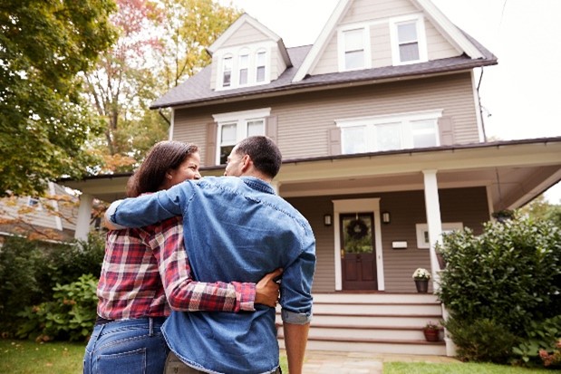 A man and woman looking at each other while hugging in front of their new home with trees to the left of them.